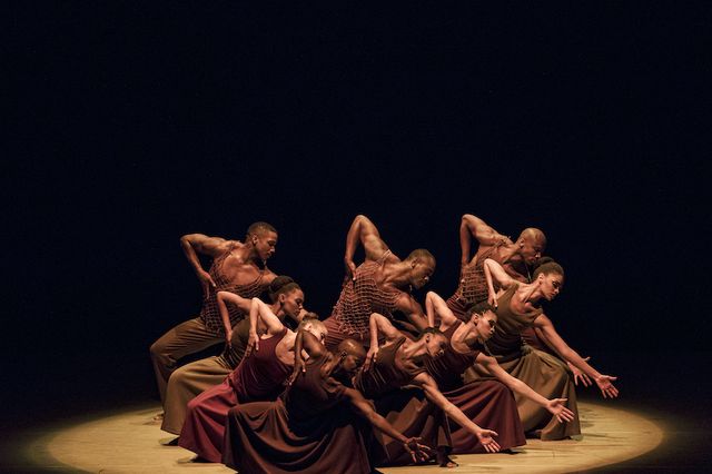 A group of dancers perform a scene from the Alvin Ailey American Dance Theater's iconic "Revelations." The dancers, all wearing brown costumes, all squat and lean to their left, reaching for the ground.